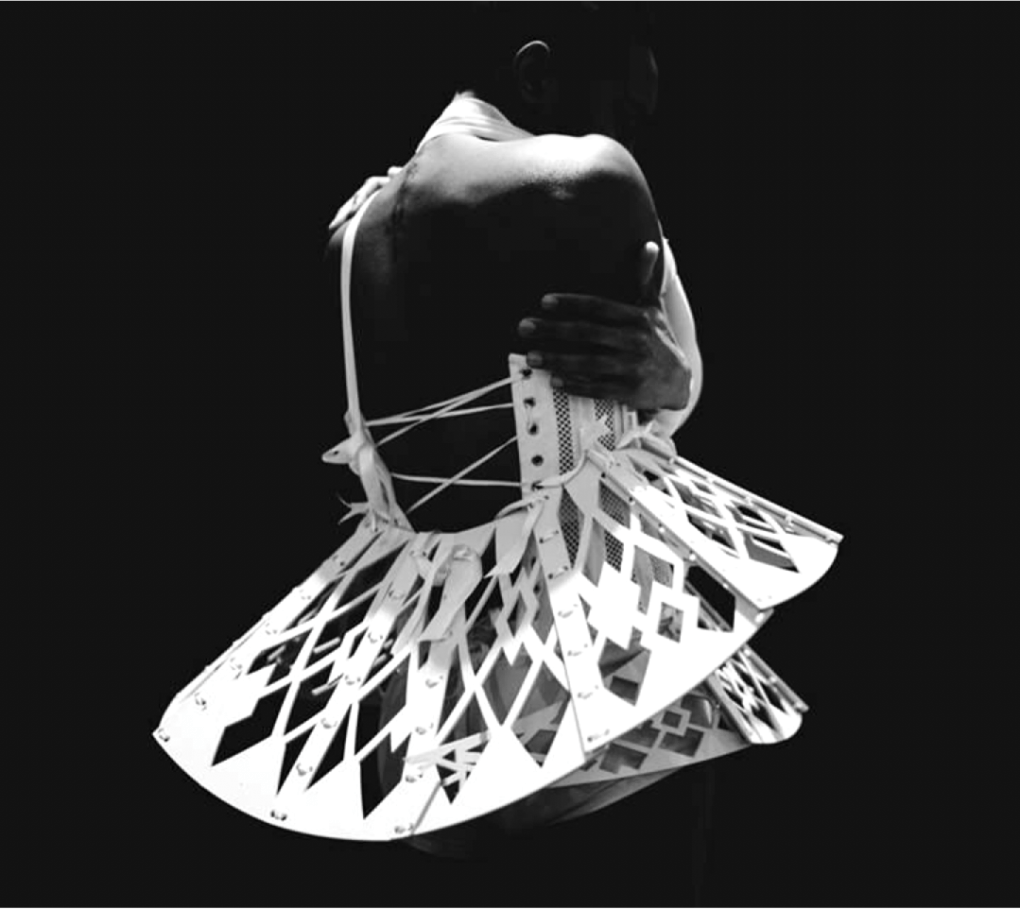 Shirtless man facing left with arms wrapped around him wearing a structural, white, wooden tutu 