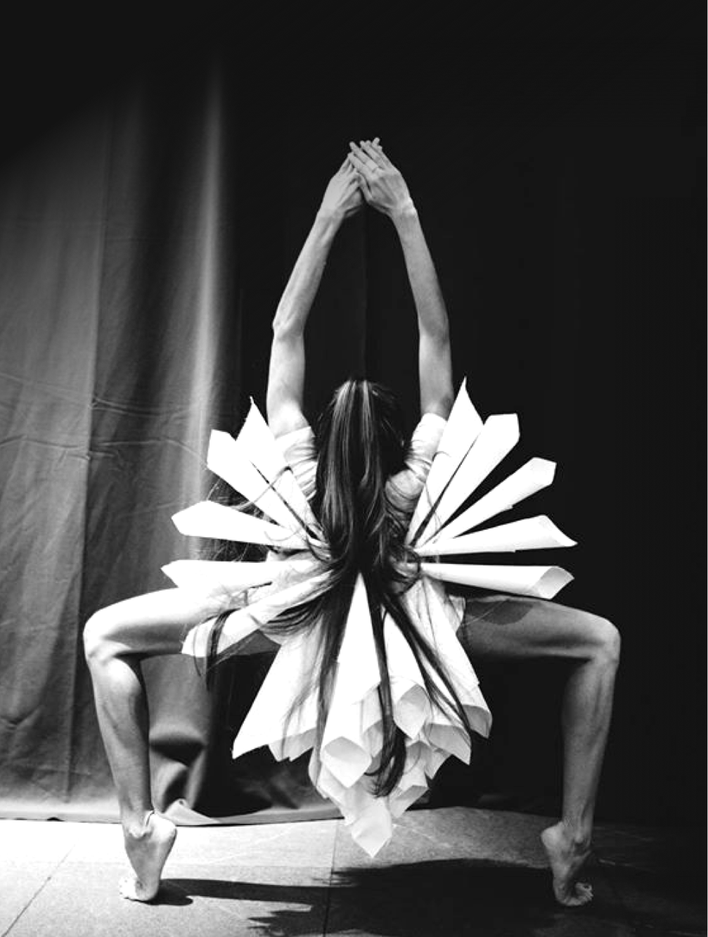 Woman with long hair facing backwards with hands clasped above her head arms in a deep bend on her toes with white tubes beaming from her center 